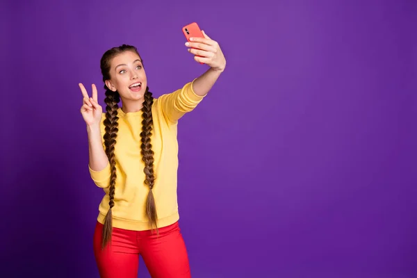 Фото красивої дівчини-підлітка довгі коси, що тримають телефон, роблять селфі, що показують символ v-знака, носять повсякденний жовтий пуловер червоних штанів ізольовані фіолетовий кольоровий фон — стокове фото