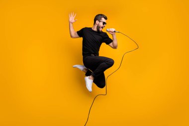 Full body profile photo of crazy hipster guy jumping high holding microphone music lover singing favorite song wear sun specs black t-shirt pants isolated yellow color background clipart