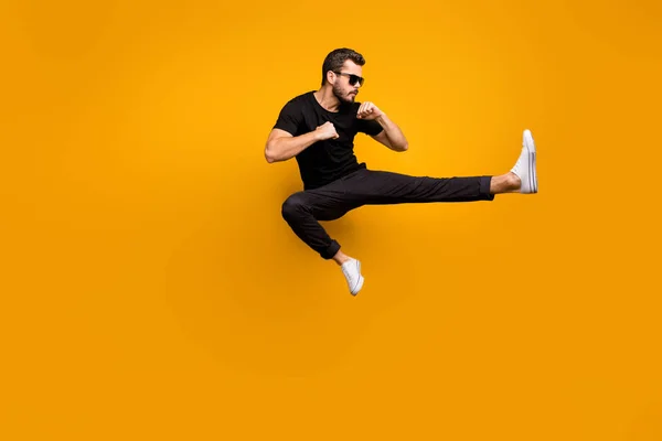 Full längd foto av stilig kille hoppar högt öva självförsvar sparkar säker ansiktsuttryck bära sol specs svart t-shirt byxor isolerad gul färg bakgrund — Stockfoto