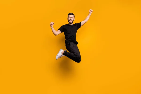 Full body foto van knappe opgewonden man springen hoog verhogen vuisten vieren voetbal team doel ondersteunen fan dragen zwarte t-shirt broek geïsoleerde gele kleur achtergrond — Stockfoto