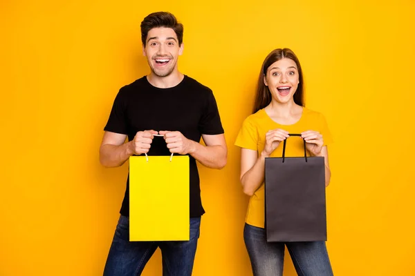 Foto de alegre positivo lindo hermosa pareja bonita celebración de paquetes de centro comercial con jeans vaqueros camiseta negra éxtasis aislado sobre fondo de color brillante — Foto de Stock