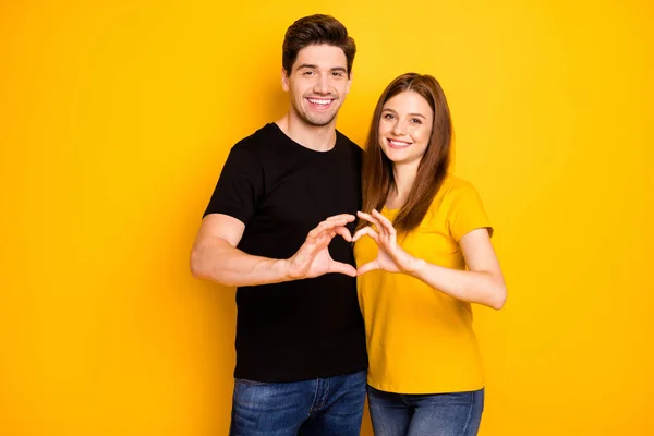 Photo of wesoły uroczy słodkie słodkie ładna para kształtowanie serca z palcami symbolizujące miłość w czarnym t-shirt dżinsy izolowane żółty żywy kolor tła — Zdjęcie stockowe