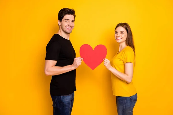 Foto de alegre bonito casal bonito compartilhando tymbol de amor grande coração vermelho uns com os outros em preto t-shirt jeans jeans isolado sobre vibrante cor amarela brilhante fundo — Fotografia de Stock