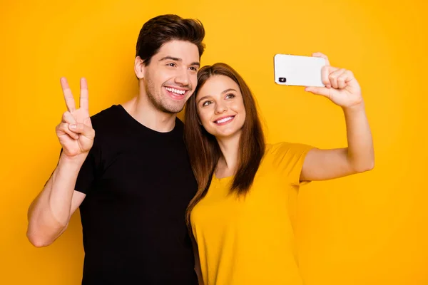 Foto de namorado e namorada sorrindo alegremente viajando ao redor do mundo mostrando v-sinal tomando selfie vestindo t-shirt preta isolado cor amarela vívida fundo — Fotografia de Stock