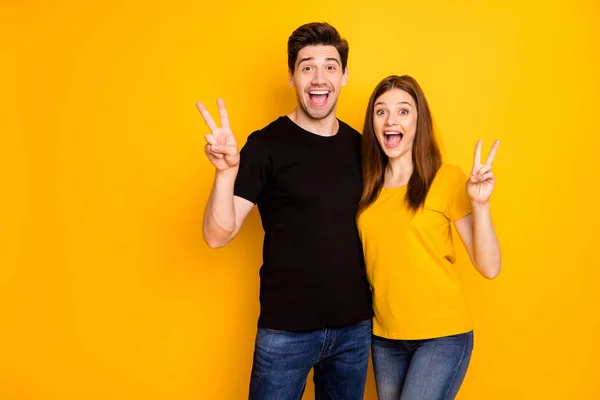 Foto de alegre y fascinante hermosa pareja con jeans vaqueros camiseta negra que muestra doble signo v con expresiones faciales emocionadas aisladas sobre un fondo amarillo de color vivo —  Fotos de Stock