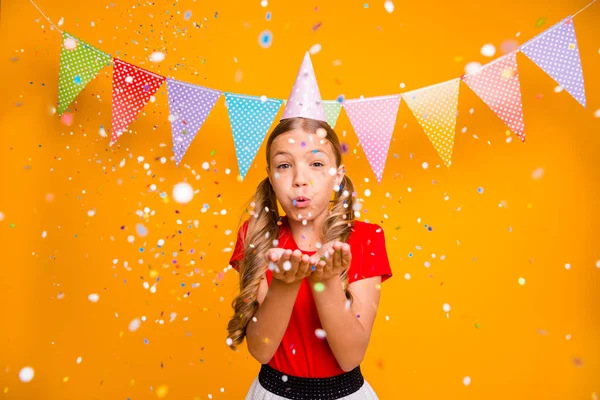 Foto van vrij grappig dametje verjaardag evenement blazen handen confetti vallen kleurrijke vlaggen opknoping muur dragen papieren hoed casual rood wit jurk geïsoleerde gele kleur achtergrond — Stockfoto