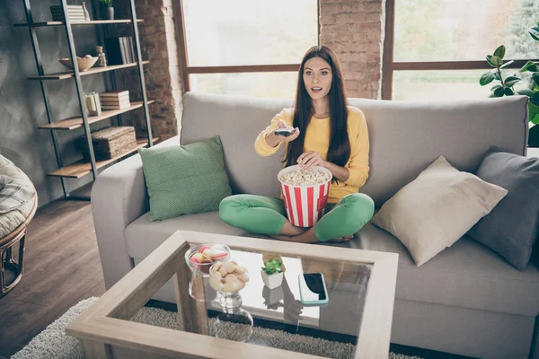 Full length photo of amazed interested girl sit divan crossed legs have free time watch film switch channels eat pop corn form big box in doors — Zdjęcie stockowe