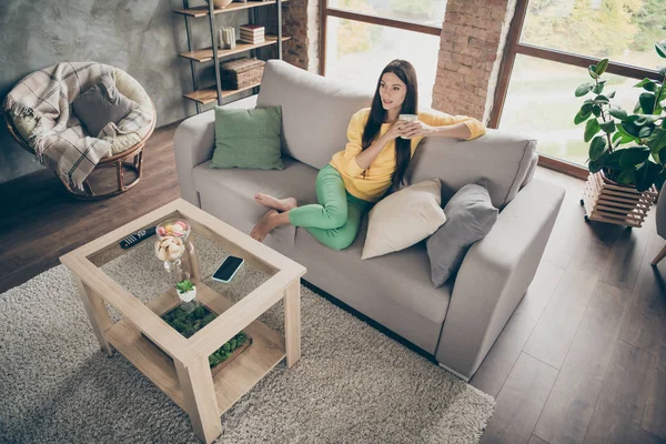 Oben über dem Hochwinkel Foto von interessierten Mädchen sitzen Diwan Uhr Film halten Tasse mit heißem Kaffee tragen gelben Pullover grüne Hosen in großen Wohnung drinnen — Stockfoto