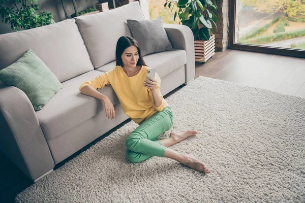 Top acima da foto de alto ângulo de menina alegre positiva sentar tapete chão uso descalço smartphone ler rede social notícias desgaste amarelo jumper calças verdes em casa dentro de casa — Fotografia de Stock