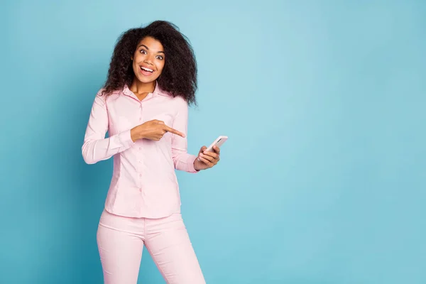 Foto de incrível ondulado escuro pele senhora segurando as mãos do telefone indicando dedo na tela venda telefone inteligente preço desgaste rosa camisa calças isolado azul cor fundo — Fotografia de Stock