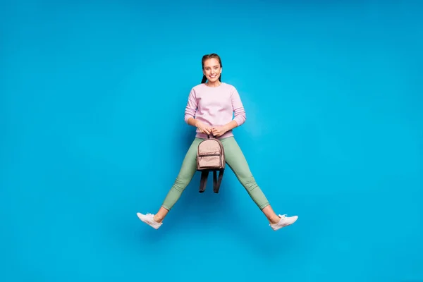 Фотографія повної довжини досить божевільної леді довгі коси, що стрибають високо готові для першого навчального дня тримайте рюкзак носити випадковий рожевий пуловер зелені штани ізольовані синій колір фону — стокове фото
