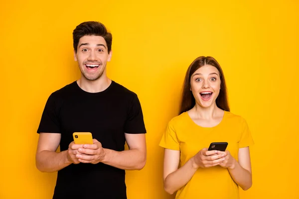 Foto de dos estudiantes locos chico dama sosteniendo teléfonos inteligentes escribiendo leyendo buenas noticias famoso influencer blog boca abierta desgaste casual negro camisetas aisladas color amarillo fondo —  Fotos de Stock