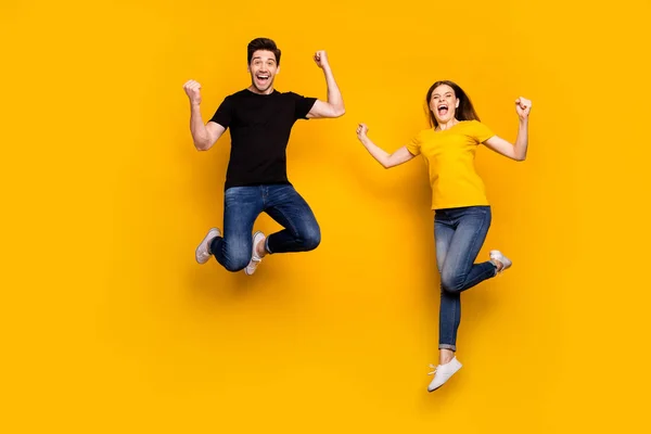 Corpo inteiro foto de cara louco senhora casal pulando alto celebrando primeiro lugar vitória competição triunfante desgaste casual jeans preto camisetas isolado cor amarela fundo — Fotografia de Stock