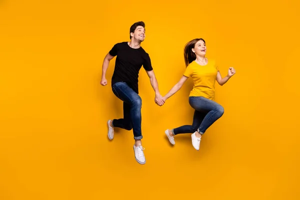 Полноразмерное фото спортивного парня и женской пары прыгающих высоко держась за руки пары участников марафона скорости носить повседневные джинсы футболки изолированный желтый цвет фона — стоковое фото