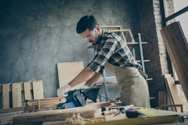 Zijprofiel foto van serieuze, zelfverzekerde man die hout verwerkt met schuurmachine met oogbescherming in geruite overhemdhandschoenen — Stockfoto