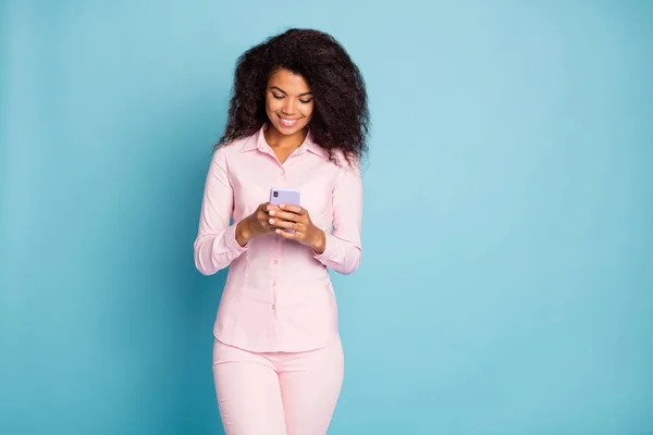 Foto di incredibile ondulato scuro pelle signora tenuta telefono controllo commenti piace seguaci repost indossare rosa camicia pantaloni isolato blu colore sfondo — Foto Stock