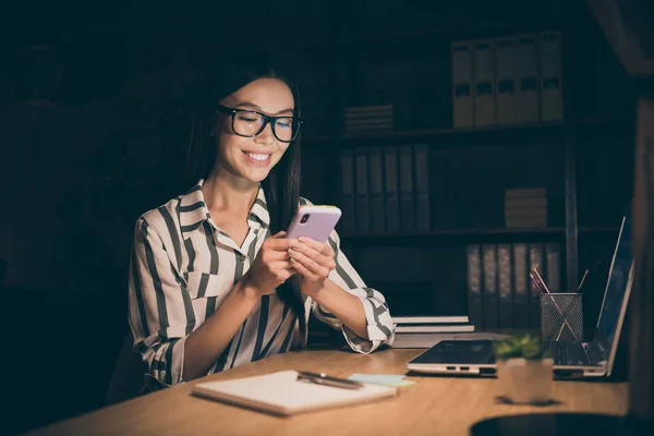 Foto de mujer de negocios bastante chino sosteniendo sms lectura de teléfono trabajando hasta tarde en la noche tienen poco descanso toothy sonriente silla de sentarse usar camisa rayada oficina moderna oscura — Foto de Stock