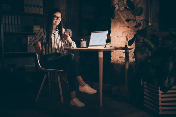 Foto di bella asiatico business signora guarda notebook tavolo tenendo telefono lettura sms lavoro fino a tarda notte hanno poco pausa sedersi sedia usura camicia a righe scuro moderno ufficio — Foto Stock