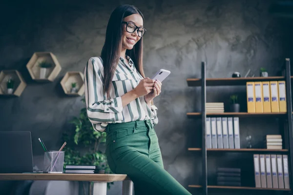 Foto de alegre jefe positivo de la corporación que descansa pasando el tiempo libre en las redes sociales sonriendo con dientes apoyados en el escritorio en gafas pantalones verdes — Foto de Stock