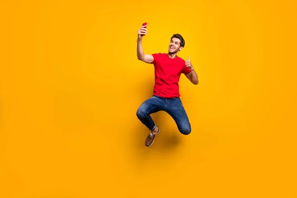 Foto em tamanho completo de cara cabelo marrom alegre desfrutar de férias de primavera salto fazer selfie mostrar gesto de polegar para cima recomendar viagem desgaste vermelho t-shirt jeans jeans isolado cor amarela fundo — Fotografia de Stock