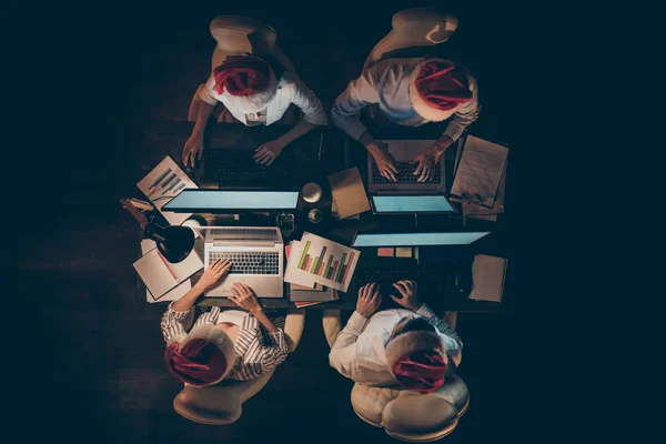 Alto ángulo por encima de la vista de la foto de cuatro personas de negocios que trabajan horas extras la noche del año nuevo buscando monitores portátiles usan gorras de santa formalwear oficina oscura en el interior —  Fotos de Stock