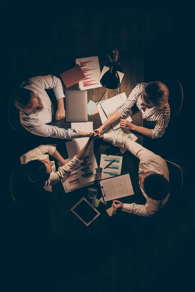 Wysoki kąt powyżej widok pionowe zdjęcie czterech osób kolegów siedzi stół pracy do późna noc trzymając stosu ręce wyrażające solidarność związkustrój formalnywewnątrz — Zdjęcie stockowe