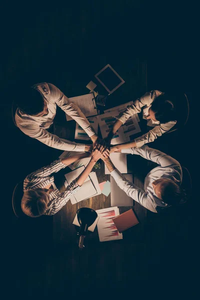 Ângulo alto acima ver foto vertical de quatro parceiros de negócios mesa sentada trabalhando juntos tarde da noite segurando braços pilha união negócio uma alma equipe formalwear lâmpada luz dentro de casa — Fotografia de Stock