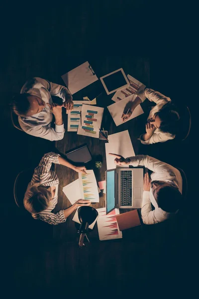 Wysoki kąt powyżej widok pionowe zdjęcie czterech partnerów biznesowych siedzi naprzeciwko stołu razem do późnej nocy omawiając nowe schematy startup szczegóły formalwear lamp światło wewnątrz — Zdjęcie stockowe