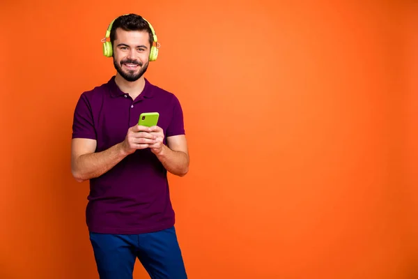 Foto de un hombre atractivo alegre y guapo sonriendo toothily sosteniendo el teléfono con auriculares inalámbricos en la cabeza en pantalones azules aislados fondo de color vibrante — Foto de Stock
