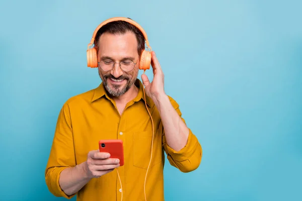 Porträt eines fröhlichen Mannes, der sein Smartphone benutzt, um Songs zu suchen, die er von seinem modernen Headset hören möchte, trägt stylische Kleidung isoliert über blauem Hintergrund — Stockfoto