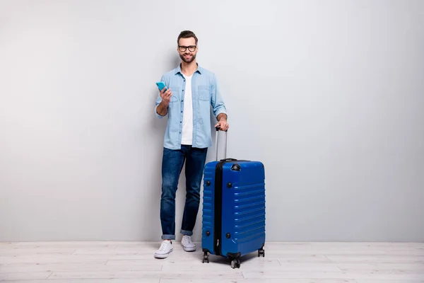 Foto de cuerpo entero del chico hipster guapo sosteniendo teléfono bolsa de turista grande tienen visa de estudiante europa viajero desgaste especificaciones traje de mezclilla aislado color gris fondo — Foto de Stock