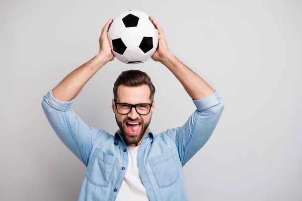 Фото сумасшедшего красивого парня, поднимающего белый черный футбольный мяч над головой кричать поддержки командный футбольный матч носить спецификации повседневной джинсовой одежды изолированный серый цвет фона — стоковое фото