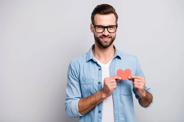 Foto de atractivo chico guapo sosteniendo poco corazón de papel rojo lleno de alegría simbolizando cardiología cuidado de la salud desgaste especificaciones traje de mezclilla casual aislado fondo de color gris — Foto de Stock