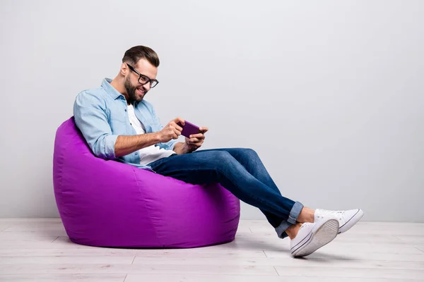 Pleine longueur profil photo de gars positif assis fauteuil violet confortable tenant téléphone jouer nouveau jeu usure spécifications casual tenue denim isolé fond de couleur grise — Photo