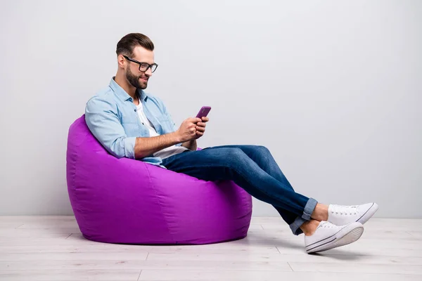 Full length profile photo of positive guy sitting comfy violet armchair holding telephone chatting friends wear specs casual denim outfit isolated grey color background — Stock Photo, Image