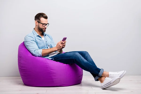 Foto de perfil em tamanho completo de cara louco sentado confortável poltrona violeta macia segurando telefone bate-papo colegas usam especificações roupa de ganga casual isolado fundo cor cinza — Fotografia de Stock