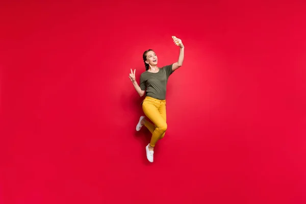 Full body foto di pazzo divertente signora salto alto facendo selfie che mostrano v-segno simbolo allegro umore indossare casual pantaloni gialli verde t-shirt isolato colore rosso sfondo — Foto Stock