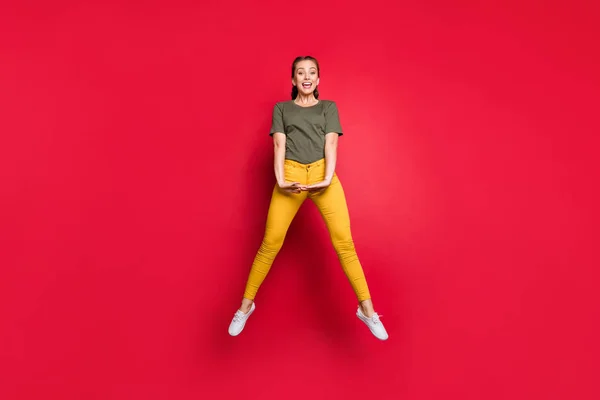 Full size foto van opgewonden duizendjarige dame springen hoog besteden vrije tijd vrolijke stemming vrolijke dragen casual gele broek groen t-shirt geïsoleerde rode kleur achtergrond — Stockfoto