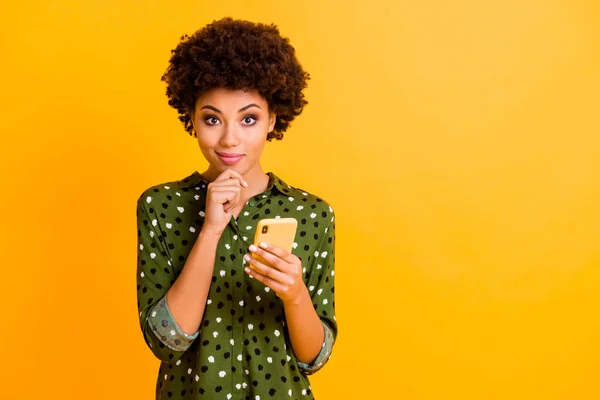 Φωτογραφία από όμορφο σκούρο δέρμα σγουρά κυρία κρατήστε τα χέρια του τηλεφώνου ενδιαφερόμενο χέρι στο πηγούνι έχουν δημιουργική μετά κείμενο ιδέα φορούν πράσινο διάστικτο πουκάμισο απομονωμένο κίτρινο χρώμα φόντο — Φωτογραφία Αρχείου