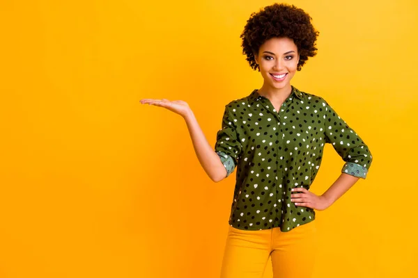Φωτογραφία από όμορφο σκούρο δέρμα κυματιστό κυρία κρατώντας καινοτόμο προϊόν στην ανοιχτή παλάμη κενό χώρο διευθυντής πωλήσεων φορούν πράσινο πουκαμίσα παντελόνι μπλούζα απομονωμένο κίτρινο χρώμα φόντο — Φωτογραφία Αρχείου