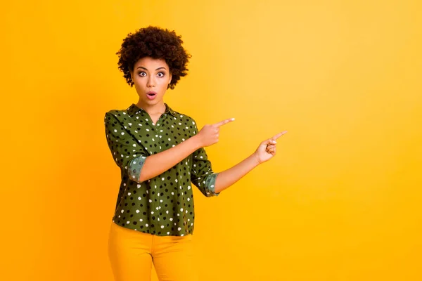 Фото смешной темно-кудрявая дама с открытым ртом указывают пальцами пустое пространство показать невероятно низкие цены носить модные зеленые брюки пунктирная рубашка изолированный желтый цвет фона — стоковое фото