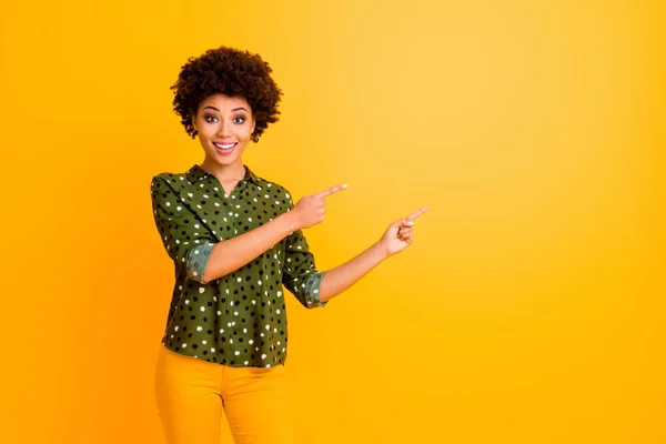 Foto de divertida señora rizada piel oscura indican los dedos lado espacio vacío aconsejando a los clientes increíbles precios bajos usan pantalones de camisa punteada verde de moda aislado color amarillo fondo — Foto de Stock