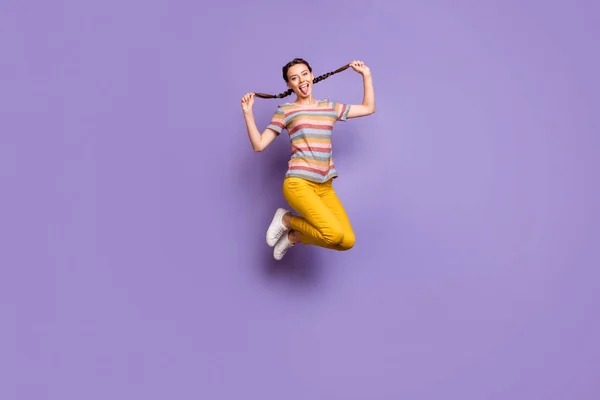 In voller Länge Foto von lustigen Dame springen Hochsommer Stimmung hält hübsche Zöpfe stecken Zunge aus Mund tragen lässig gestreift T-Shirt gelb Hose isoliert lila Farbe Hintergrund — Stockfoto