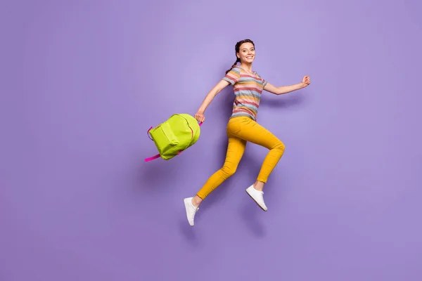 Foto de tamaño completo de la señora emocionada loca saltando alta llevar mochila verde de la escuela corriendo a casa después de las lecciones usan casual rayas camiseta pantalones amarillos aislados color púrpura fondo — Foto de Stock