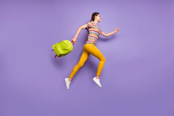 全身上下都是疯狂兴奋的女士跳高中时的照片绿色背包，下课后穿着休闲条纹T恤黄裤子，背景是紫色 — 图库照片