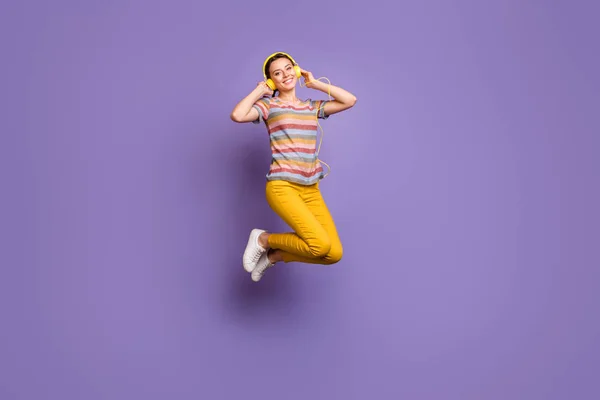 Teljes hosszúságú fotó vonzó csinos hölgy jumping magas hallgatás hűvös fülvédők ifjúsági chill party dalok viselnek alkalmi csíkos póló sárga nadrág elszigetelt lila színű háttér — Stock Fotó