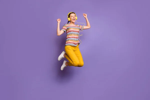 全长照片，时髦的女士跳高听着凉爽的耳罩签署疯狂的青春歌曲穿着休闲条纹T恤黄裤子孤立的紫色背景 — 图库照片