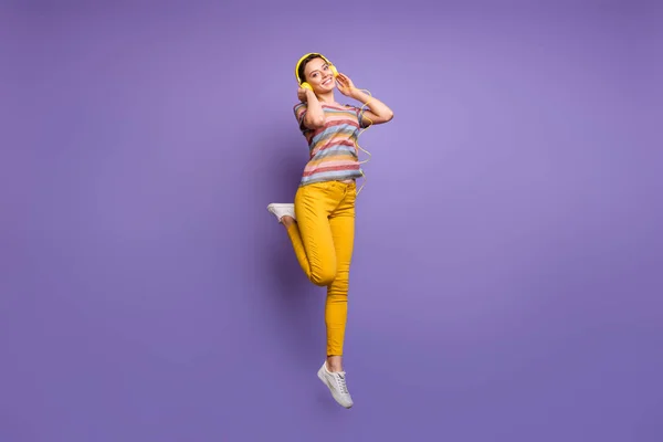 美しい女性の完全な長さ写真ジャンプ高いリスニングクールな耳フラップ若者のチルパーティーの歌はカジュアルストライプtシャツ黄色のズボン孤立紫色の背景を身に着けている — ストック写真