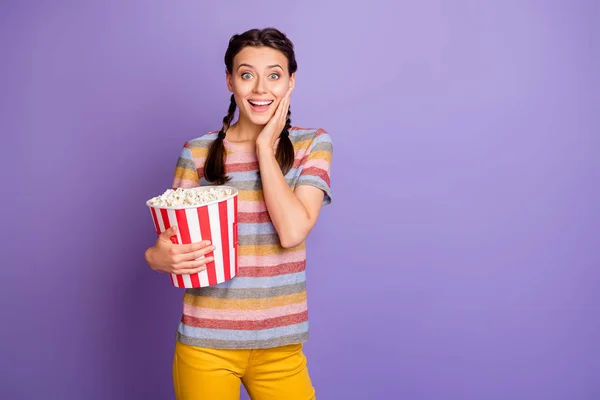 Porträtt av funky tonåring ung flicka titta på rolig film hålla stora popcorn box imponerad skrik wow omg touch hand ansikte bära gul tusenårig outfit isolerad över lila färg bakgrund — Stockfoto