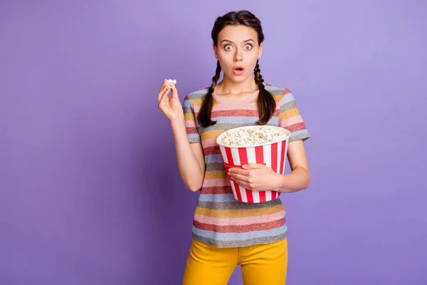 Portret van verbaasd tiener meisje kijken tv horror thriller film houden grote witte rode popcorn doos dragen casual stijl kleding geïsoleerd over violet kleur achtergrond — Stockfoto